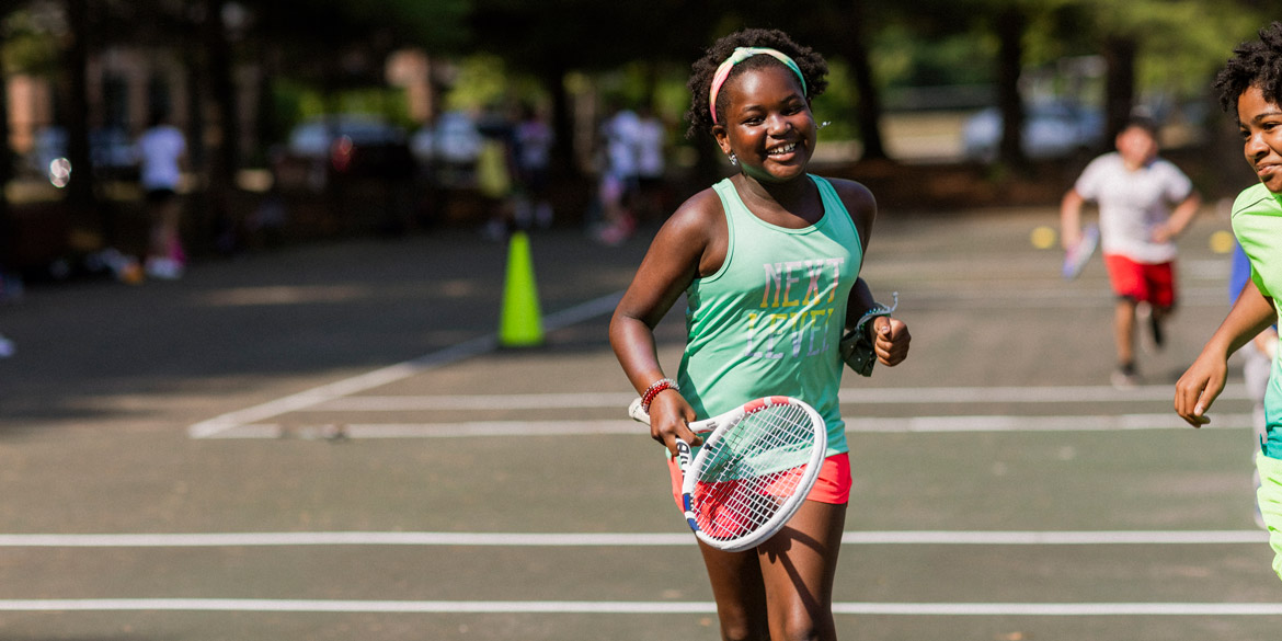 youth athlete running besides a teammate on a tennis court smiling while holding a racquet