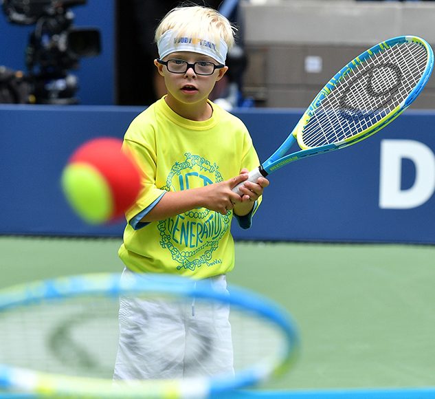 A young child in a yellow t-shirt holds a tennis racquet,. looking at a tennis ball and racquet in the foreground.