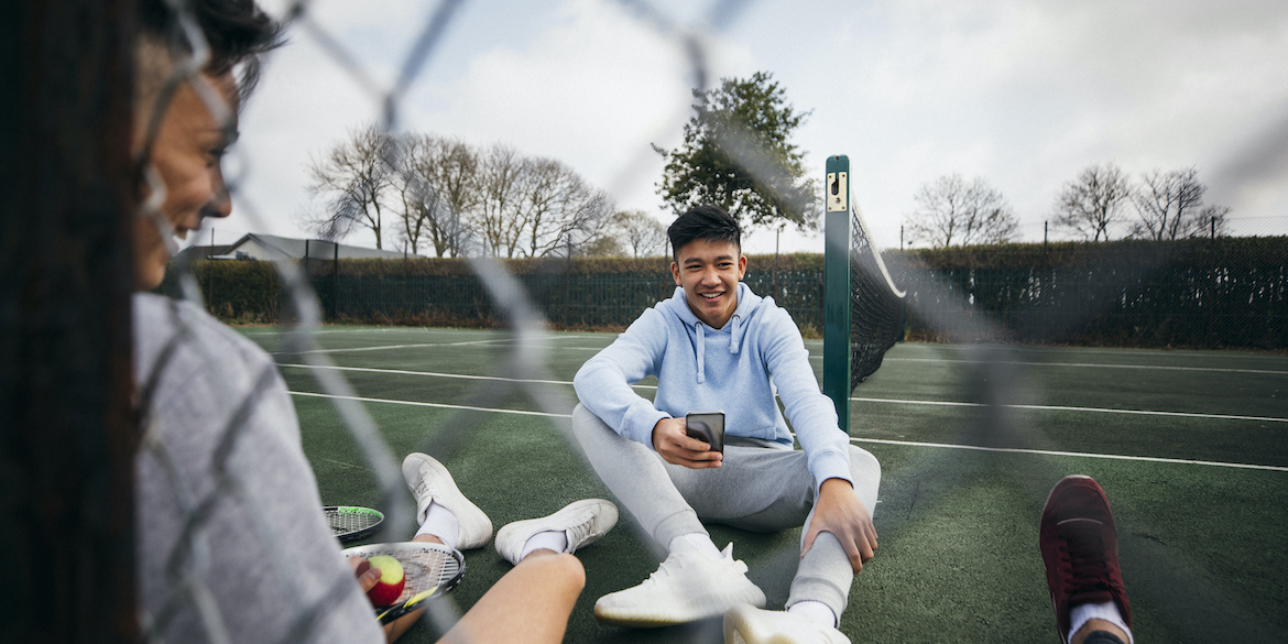 Teenage boys relaxing while sitting on a tennis court. One teenage boy is using his smart phone while using social media.