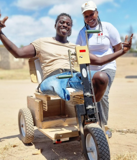 An African-American sits on a mobility cart with a big smile and hands outstretched. The individual is flanked by a lady who is also smiling.