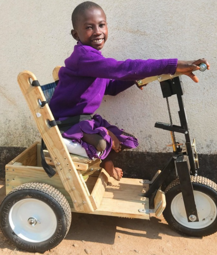 A young African-American child in a purple top smiles while sitting on a mobility cart.
